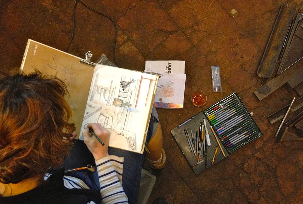 Photo prise d'en haut d'une voyageuse en train de croquer, on peut observer son carnet sur lequel elle est en train de dessiner et sa boite à crayons multicolores à sa droite